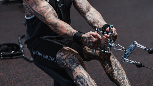 heavily tattooed individual performing a seated cable row exercise in a gym, showcasing muscular arms and legs with detailed tattoos.