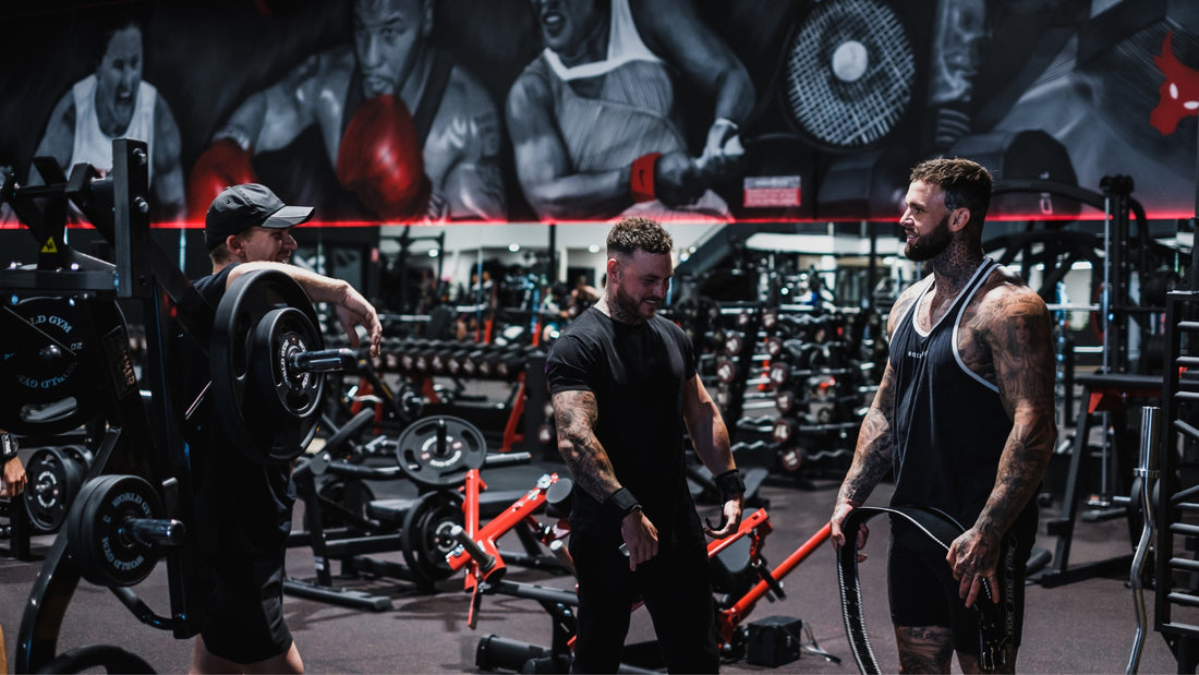 Three men are seen sharing a lighthearted moment in a gym filled with weightlifting equipment. They are muscular and tattooed, dressed in workout attire, and appear to be discussing their routines or encouraging each other.