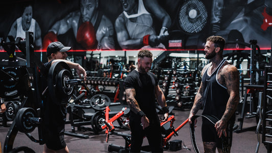 Three men are seen sharing a lighthearted moment in a gym filled with weightlifting equipment. They are muscular and tattooed, dressed in workout attire, and appear to be discussing their routines or encouraging each other.