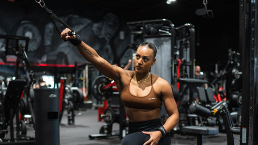 Fit woman in a brown sports bra performing a single-arm cable exercise in a dimly lit gym, focusing on strength training.