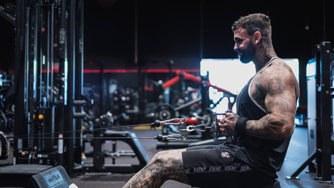 Man with tattoos performing seated cable row exercise at the gym, focusing on his back and arm muscles in a dark, intense gym environment